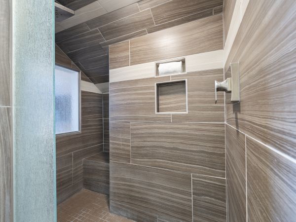 The image shows a modern tiled shower with a bench, a recessed shelf, and a frosted window, featuring gray and beige tones arranged in a contemporary pattern.