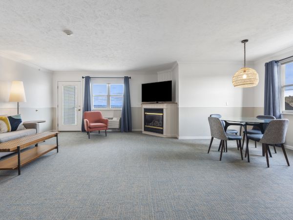 A spacious living area with a sofa, chair, wall-mounted TV, lamp, fireplace, dining table with chairs, and large windows with curtains.