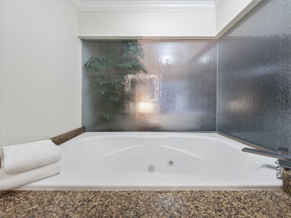 A white bathtub with a towel on the side, granite countertop, and frosted glass window with a view of a plant and lamp in the background.