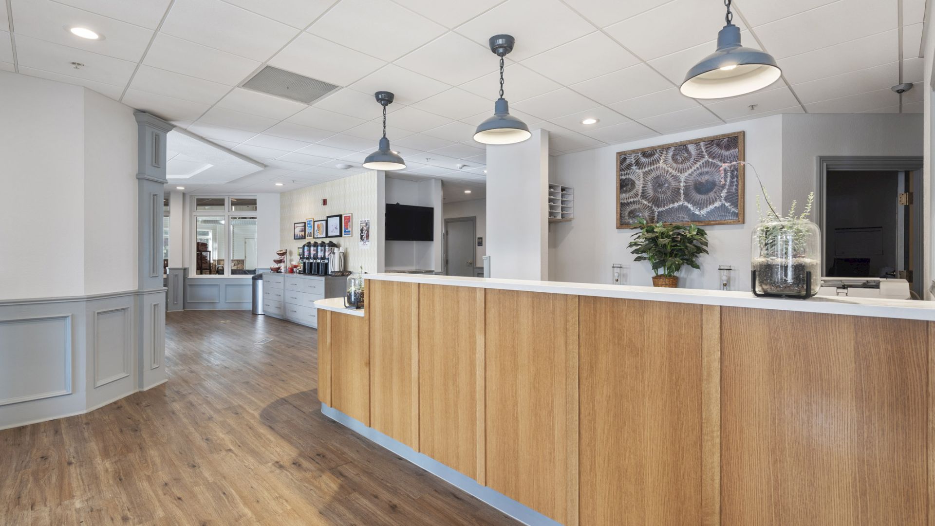 The image shows a reception area with a wooden counter, pendant lights, and a potted plant. There is a seating area in the background.