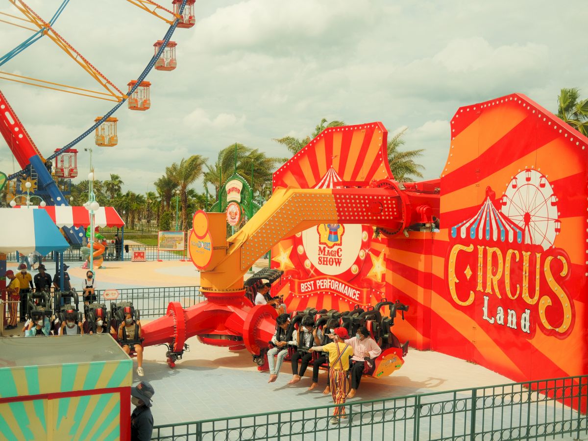 An amusement park ride with "Circus Land" signage, bright colors, and people seated and waiting for the ride to start, surrounded by other park attractions.