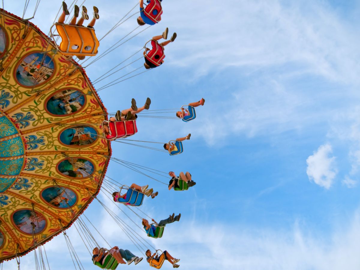 People are riding a colorful swing carousel against a blue sky. The riders are seated in chairs connected by chains, swinging outward.