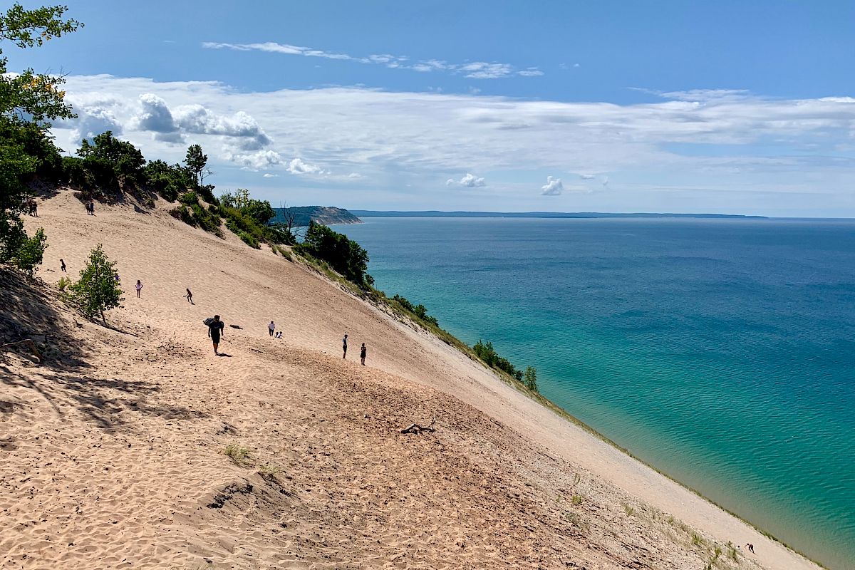 People are walking on a large sandy dune next to a blue body of water, with the sky partly cloudy and trees on the higher ground.