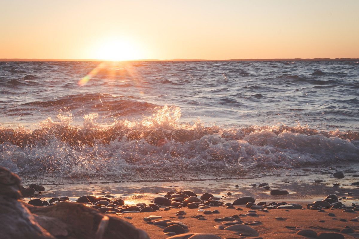 The image shows a serene beach scene with gentle waves, a pebbled shore, and a beautiful sunset casting a warm glow over the water.