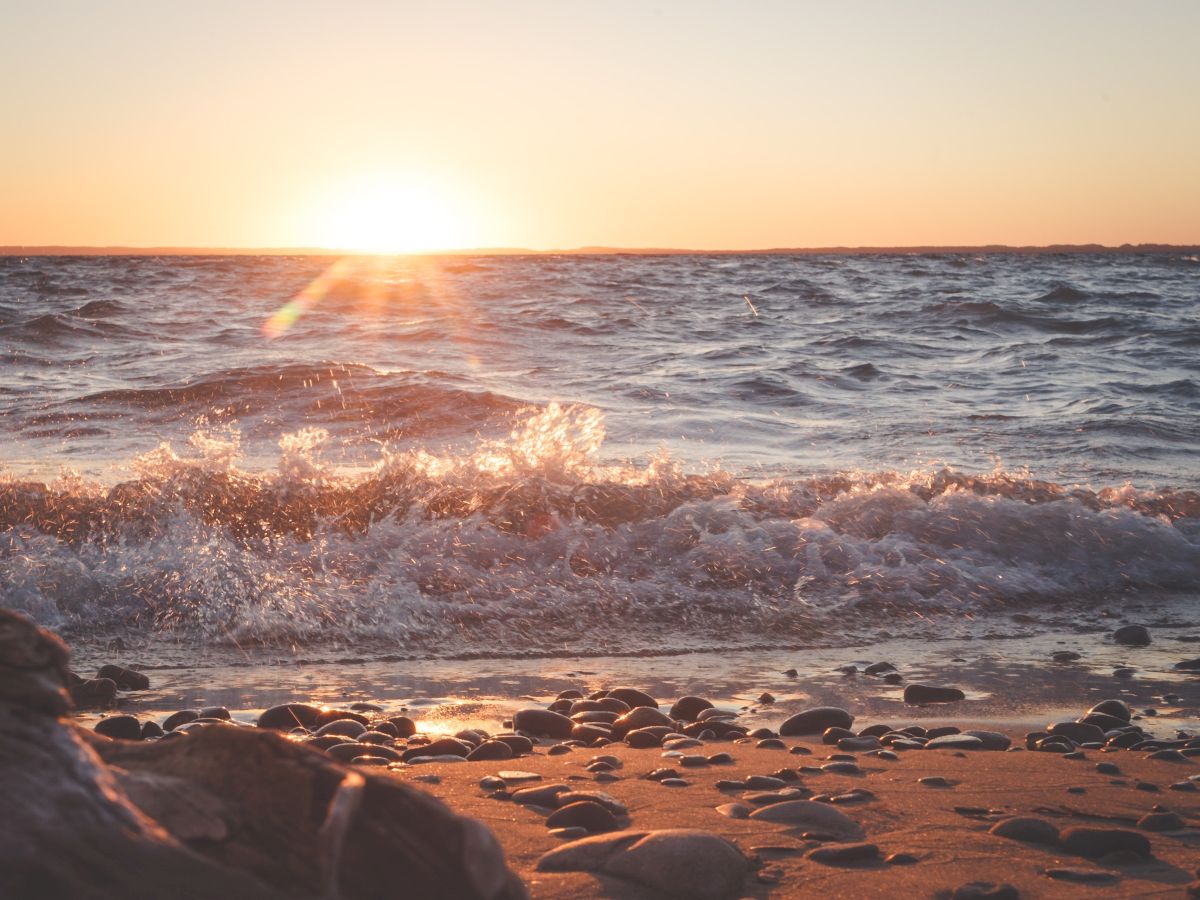 The image shows a serene beach scene with gentle waves, a pebbled shore, and a beautiful sunset casting a warm glow over the water.