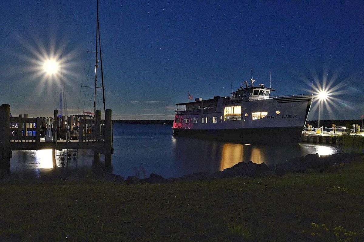 A well-lit boat docks at night with reflections shimmering on the water and starry skies above, creating a tranquil and scenic view.
