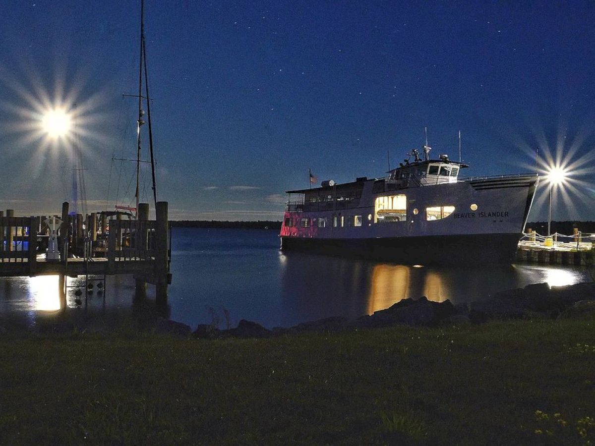 A well-lit boat docks at night with reflections shimmering on the water and starry skies above, creating a tranquil and scenic view.