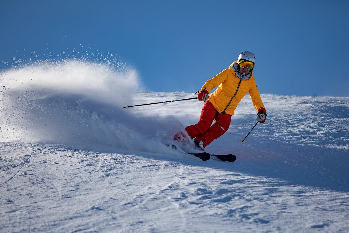A person is skiing on snow wearing a yellow jacket and red pants, creating a spray of snow as they make a turn.