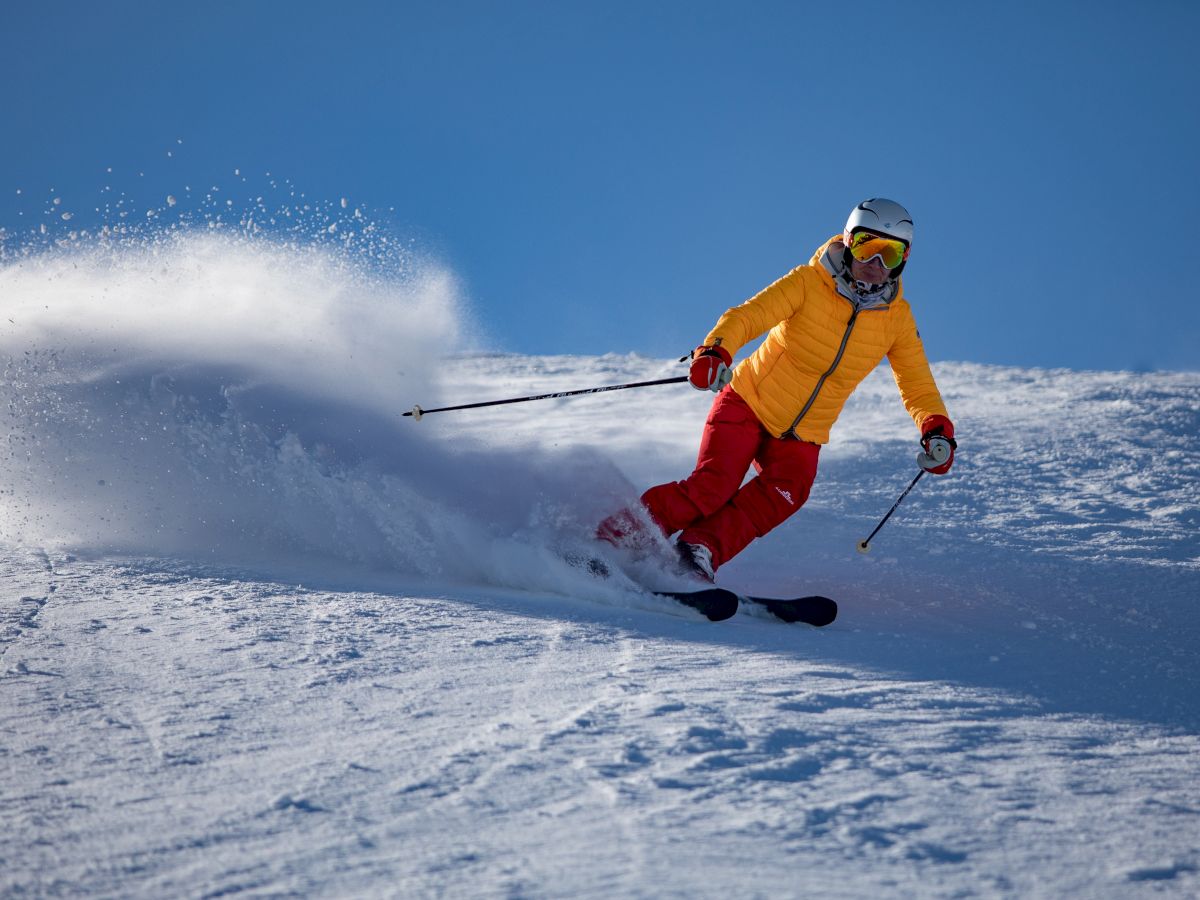 A person is skiing on snow wearing a yellow jacket and red pants, creating a spray of snow as they make a turn.