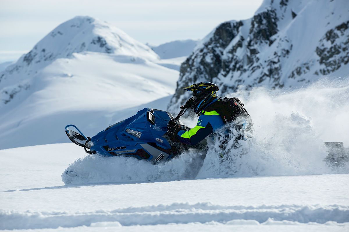 A person is riding a blue snowmobile through snowy mountains, creating a spray of snow behind them.