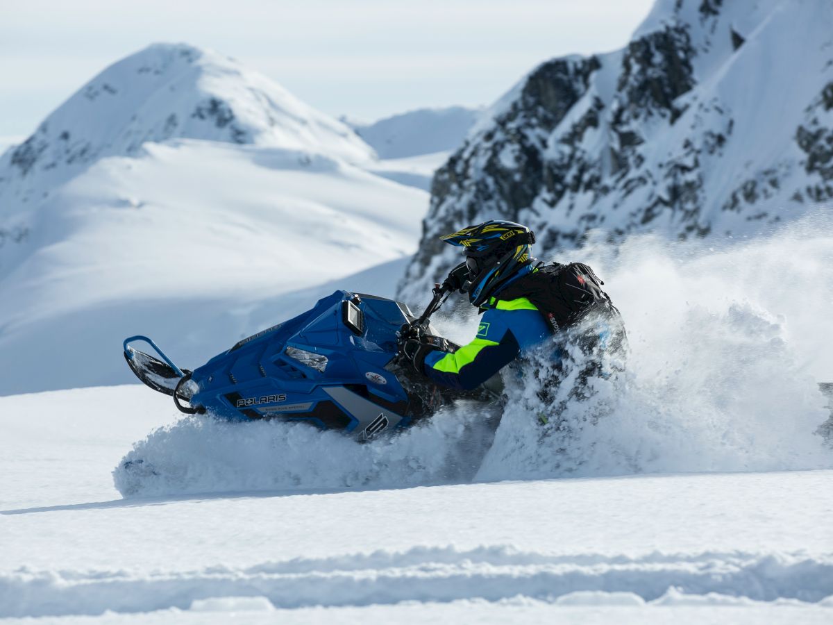 A person is riding a blue snowmobile through snowy mountains, creating a spray of snow behind them.