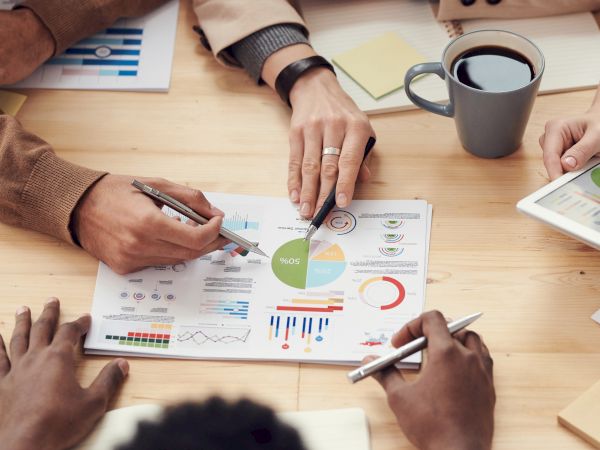 Several people analyzing charts and graphs at a meeting; there is a tablet, papers, pens, and a cup of coffee on the table.