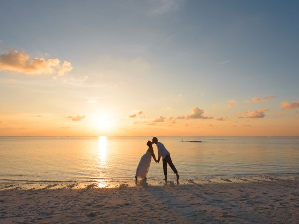 A couple is kissing on a beach at sunset with the sun setting on the horizon, surrounded by calm water and a few clouds in the sky.