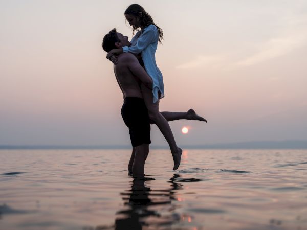 A couple wading in shallow water at sunset, the man lifting the woman as they kiss above the calm sea.