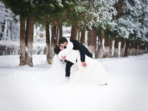 A couple, dressed in wedding attire, shares a romantic kiss in a snowy landscape surrounded by trees.