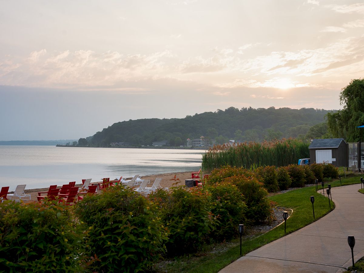 A tranquil lakeside scene at sunset with a curving walkway, red lounge chairs along the shore, and a tree-lined path beside the water.