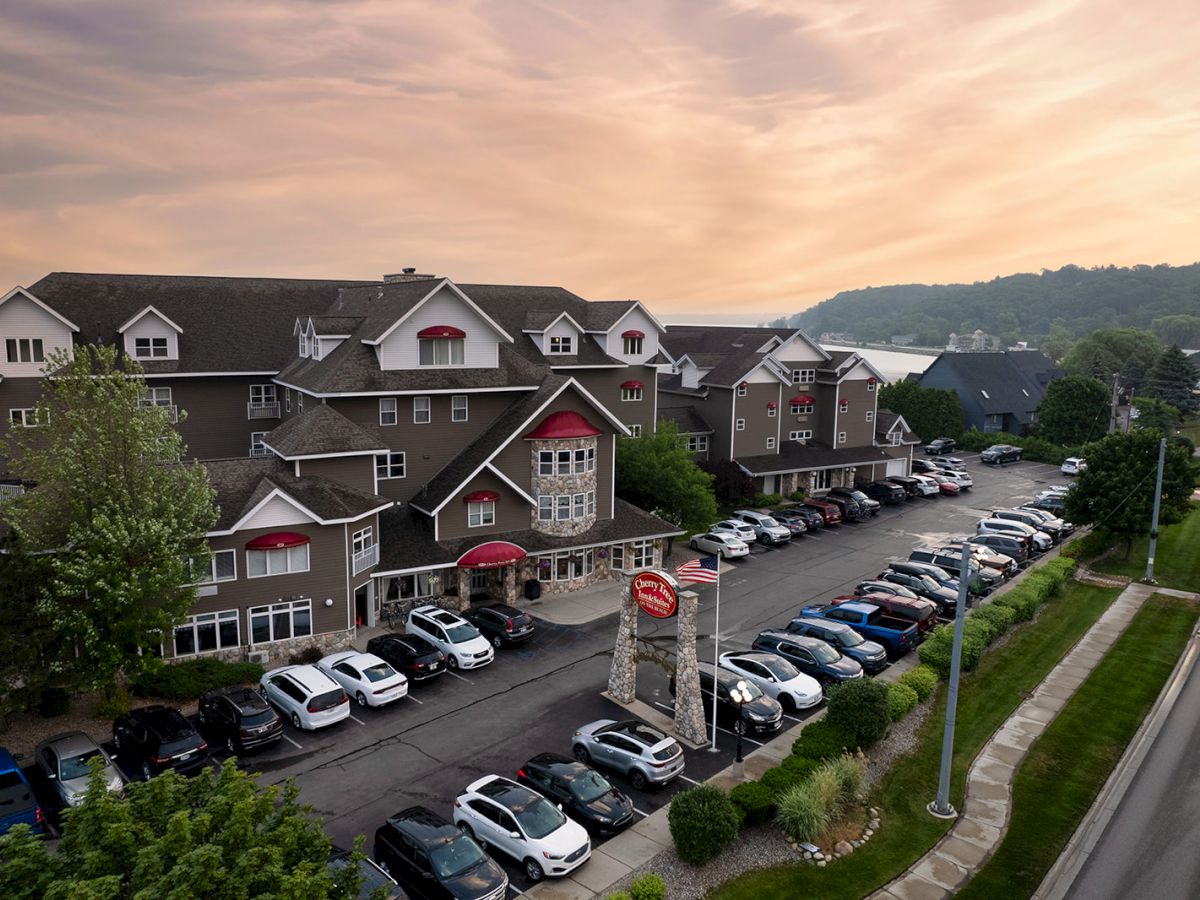 A large hotel or apartment complex with multiple gabled roofs, a long row of parked cars along the front, and a sunset sky over a hilly backdrop.