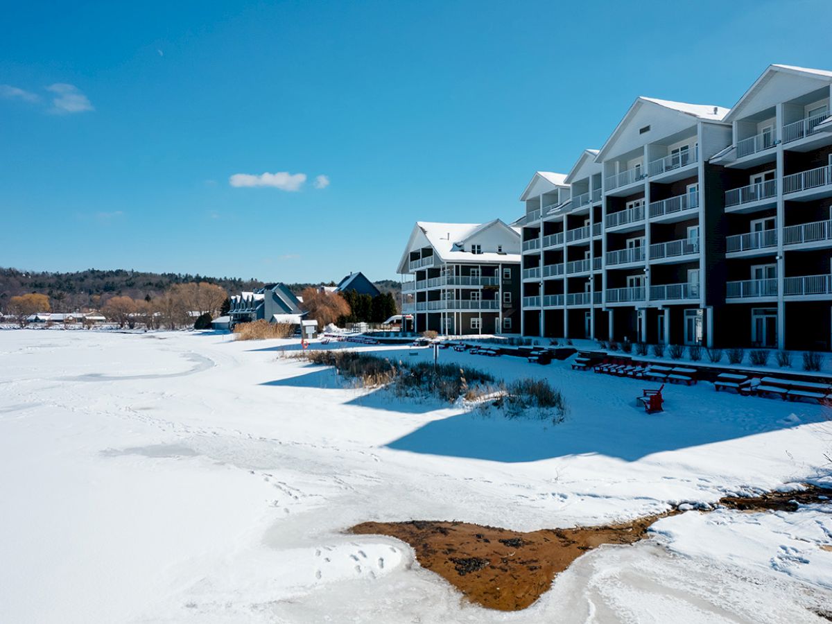 A snowy landscape with a large building featuring multiple balconies sits alongside a frozen body of water under a clear blue sky.