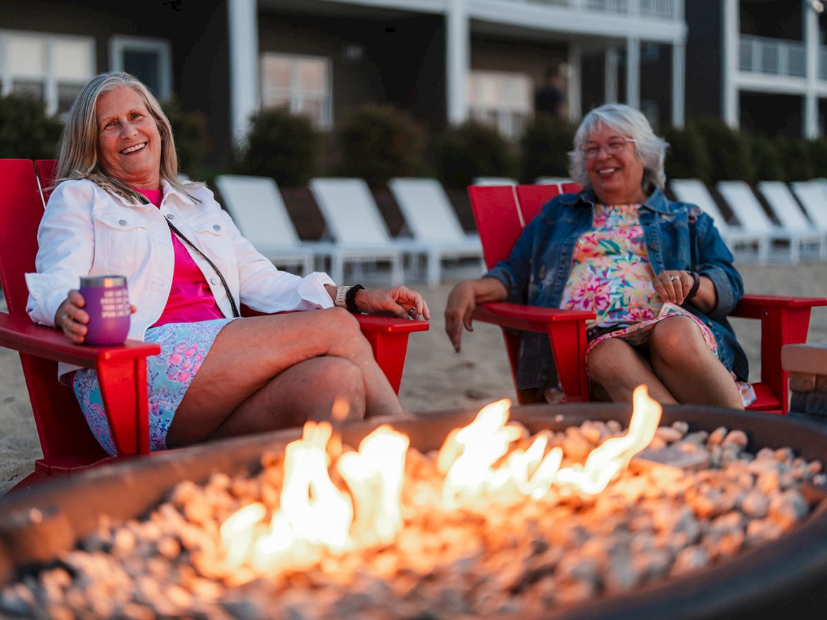 Two people sit smiling in red chairs by a fire pit on a beach, with buildings and lounge chairs in the background.