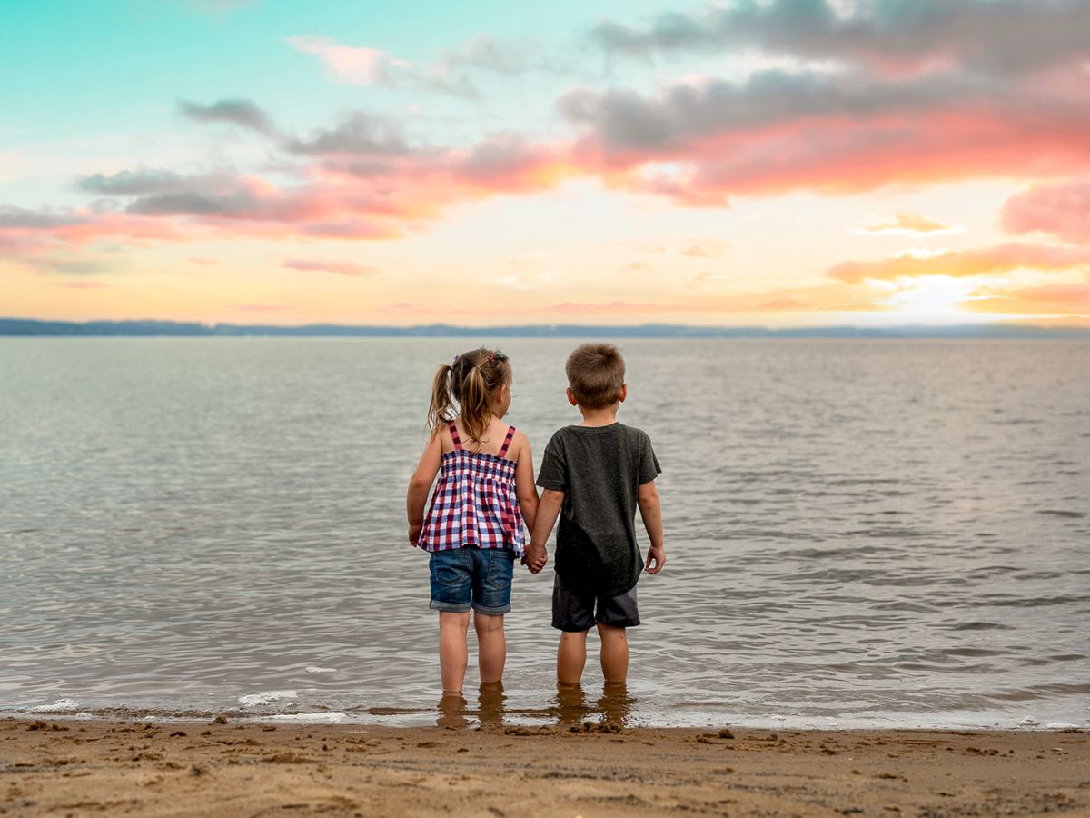 Two children holding hands, standing at the edge of a beach, looking at the water under a colorful sky during sunset.