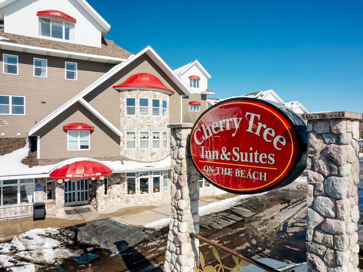 The image shows a building with a sign reading "Cherry Tree Inn & Suites Traverse City," featuring snowy surroundings under a clear blue sky.