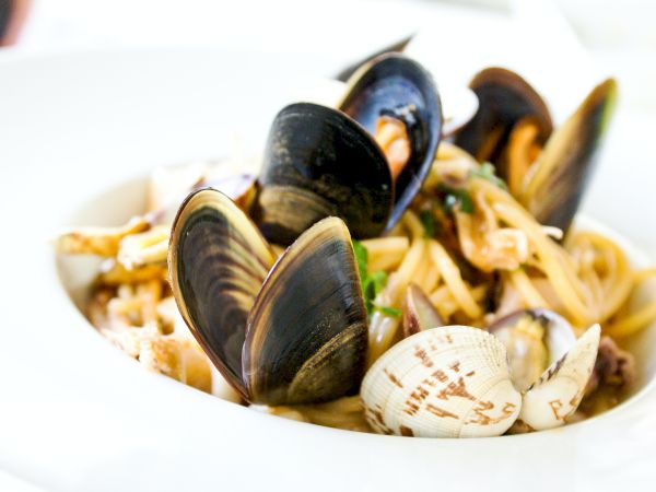 The image shows a bowl of seafood pasta, with shellfish including clams and mussels, mixed with spaghetti and garnished with herbs.