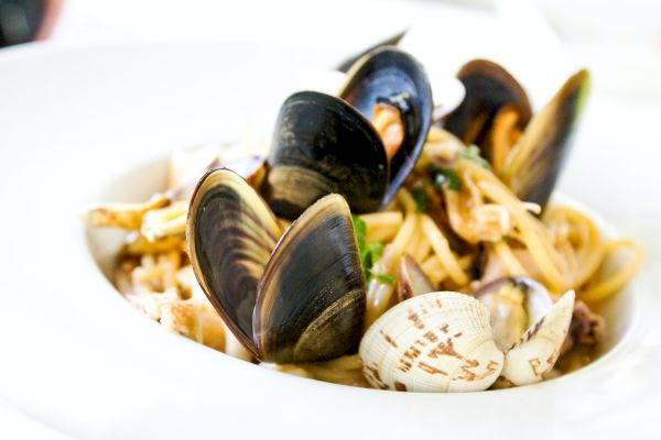 The image shows a bowl of seafood pasta, with shellfish including clams and mussels, mixed with spaghetti and garnished with herbs.