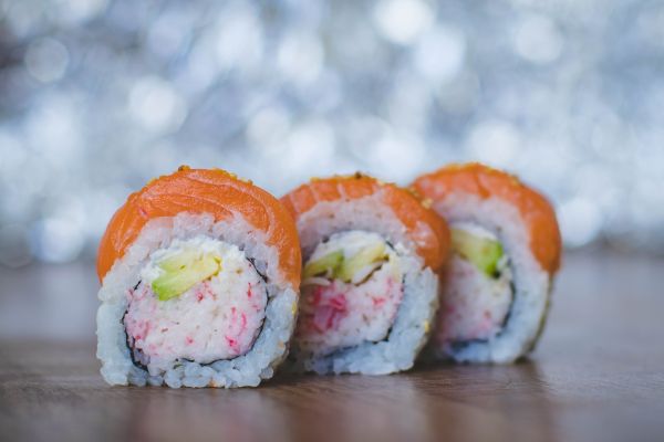 The image shows three sushi rolls with rice, avocado, and a pink filling, wrapped in seaweed and topped with salmon, on a wooden surface.