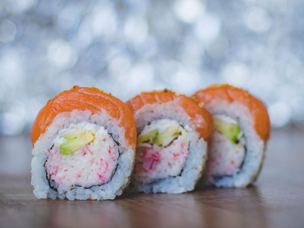 The image shows three sushi rolls with rice, avocado, and a pink filling, wrapped in seaweed and topped with salmon, on a wooden surface.