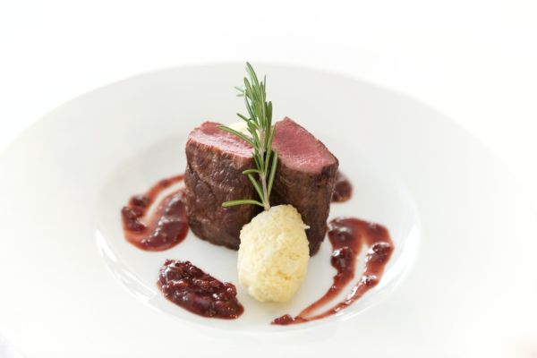 A gourmet dish featuring a piece of seared meat, garnished with mashed potatoes and a sprig of rosemary, with a berry sauce drizzled on the plate.