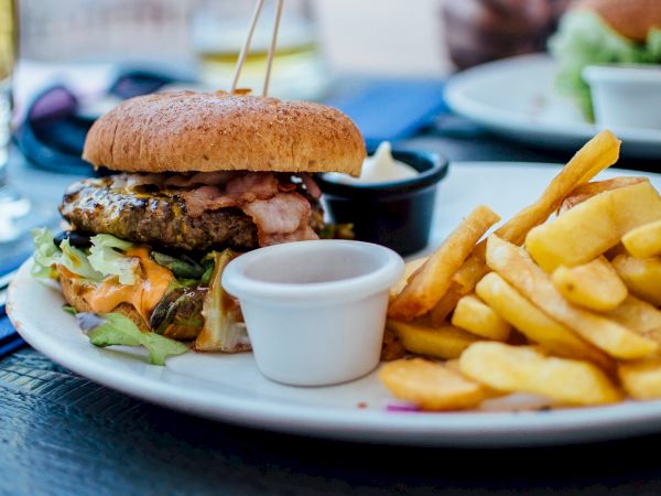 A plate of food featuring a burger with toppings, a side of fries, and dipping sauces is shown on a table.
