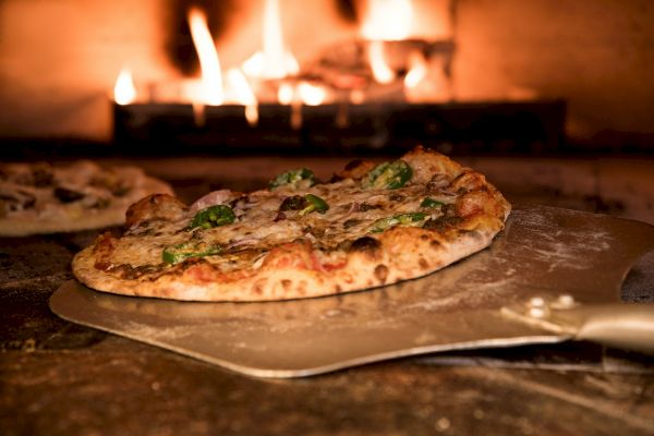 A freshly baked pizza on a pizza peel is being taken out of a wood-fired oven with flames in the background.