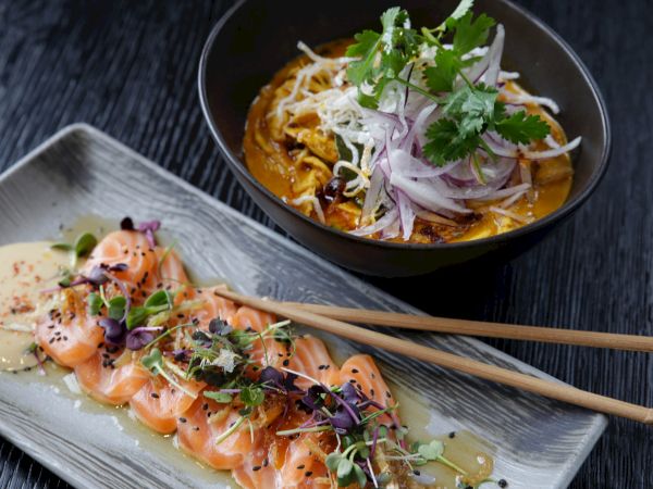 The image shows a bowl of salad with greens and a dish of sliced salmon sashimi garnished with herbs and a creamy sauce, topped with chopsticks.