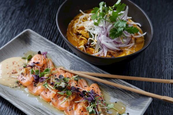 The image shows a bowl of salad with greens and a dish of sliced salmon sashimi garnished with herbs and a creamy sauce, topped with chopsticks.