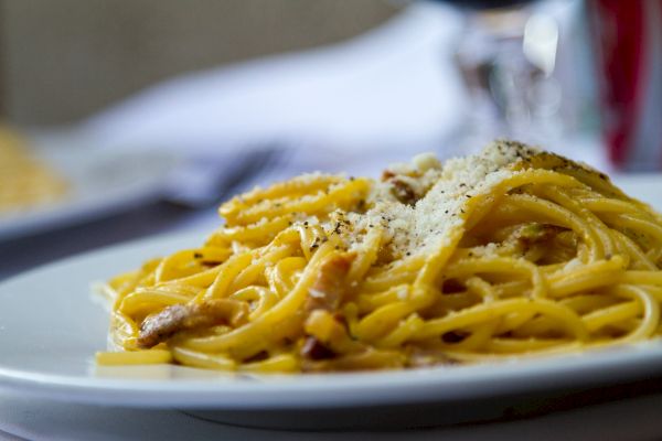 A plate of spaghetti carbonara topped with grated cheese and black pepper. The pasta dish is served on a white plate, in focus.