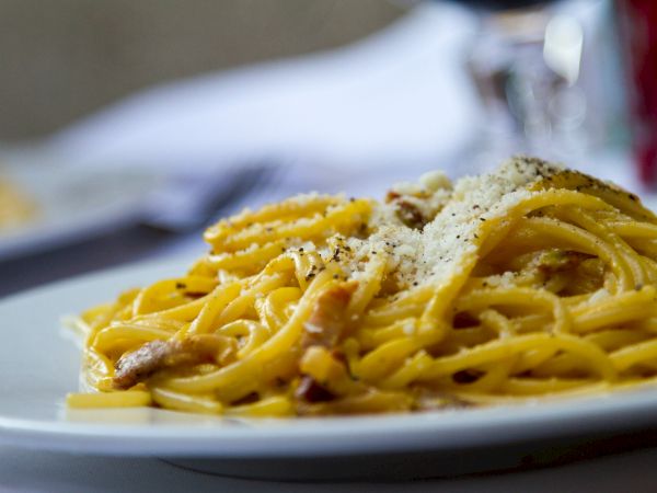 A plate of spaghetti carbonara topped with grated cheese and black pepper. The pasta dish is served on a white plate, in focus.