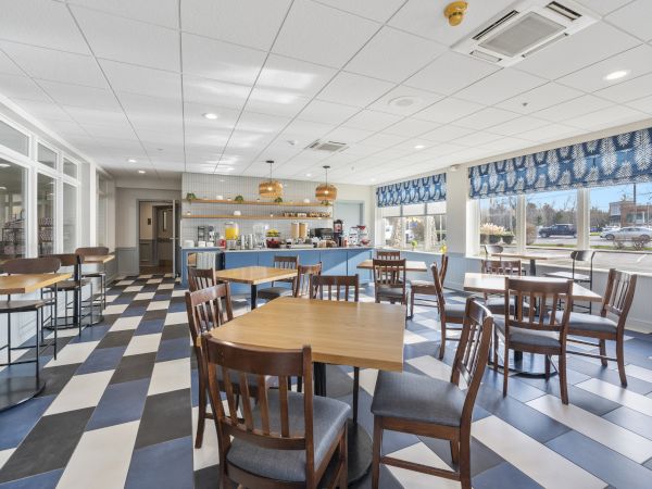 A bright, modern dining area with wooden tables and chairs, a checkered floor, large windows, and a counter with hanging lights in the background.