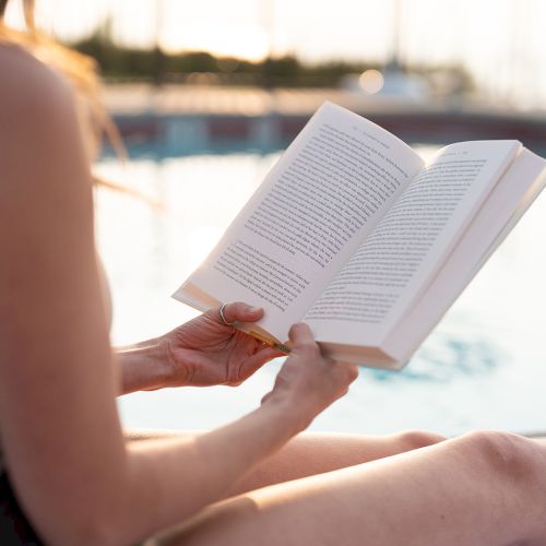 A person is relaxing by a pool, reading an open book in their hands.