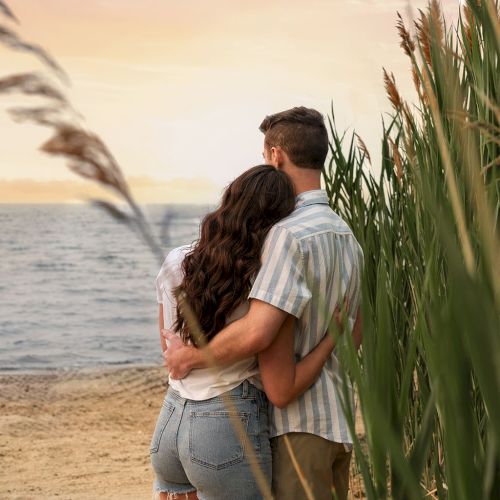Two people embrace and kiss by a beach, framed by a palm-lined path at sunset.