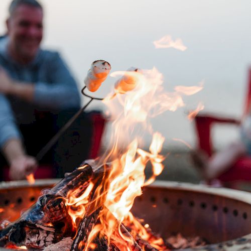 Three friends around a campfire on a beach at sunset, roasting marshmallows and chatting, relaxed and warm vibe.