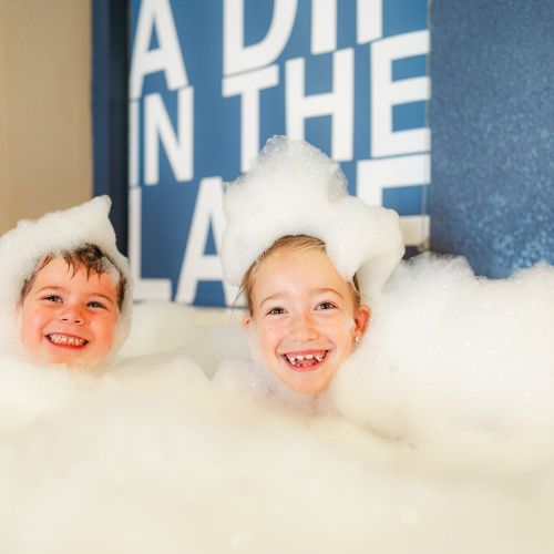 Two children are smiling while submerged in a large amount of bubbles, with part of a sign visible in the background.