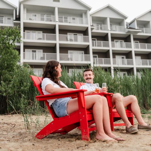 A couple sitting on red chairs with drinks, on sandy ground by tall grass, in front of a multi-story building.