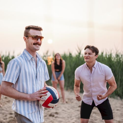 Four people on a beach, one holding a volleyball, with grass in the background. They appear to be relaxed and enjoying the moment.