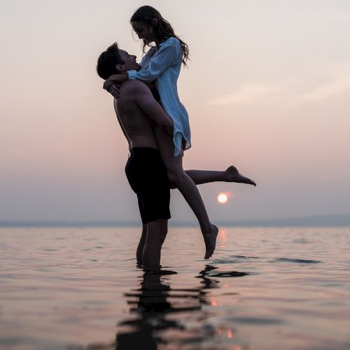 A couple is in the water at sunset; the man lifts the woman while they embrace, reflecting on the calm surface.