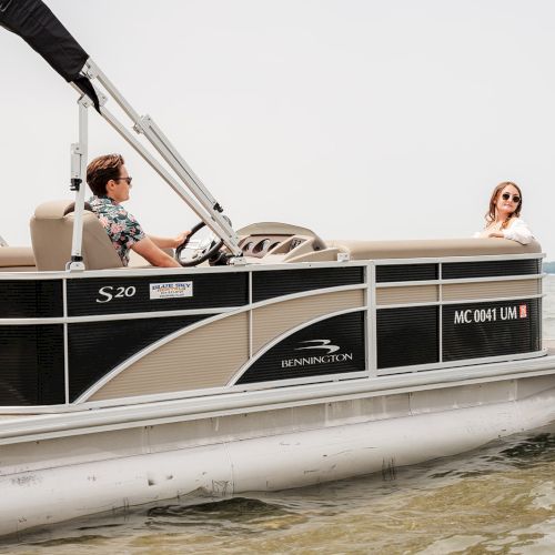 Two people are on a pontoon boat in the water, with one person steering and the other sitting on the side.