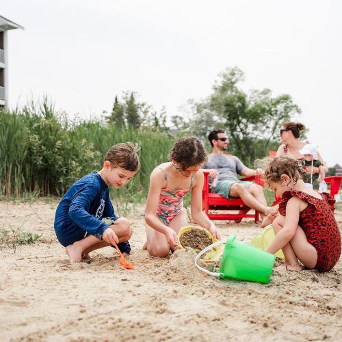 A group of kids sits and plays with a sandcastle on a sunny day near a fence and trees, smiles and enjoys outdoor fun.