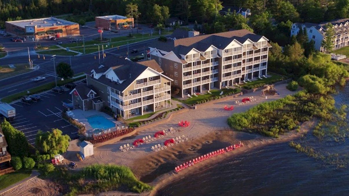 A beachfront hotel with a pool, beach chairs, and umbrellas. Adjacent road and buildings in the background, surrounded by greenery, ending the sentence.