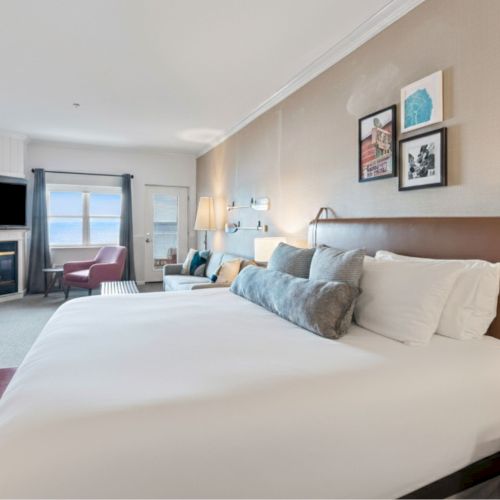 A spacious hotel room with a large bed, wall art, a fireplace, seating area, and a window with curtains in a light, modern decor style.