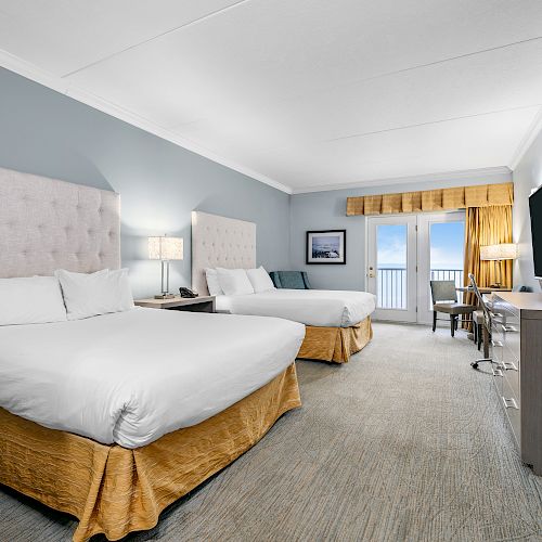 A hotel room with two large beds, a TV, a desk, and a view of a balcony overlooking the sea. Beige and gray color scheme with modern decor.