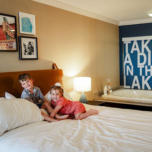 Two children on a bed in a cozy room with framed art on the wall and a jacuzzi tub in the background that has a sign saying "TAKE A DIP IN THE LAKE."